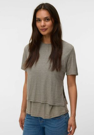 Femme aux longs cheveux foncés portant un haut gris côtelé à manches courtes et superposé ainsi qu'un jean bleu sur un fond clair uni.
