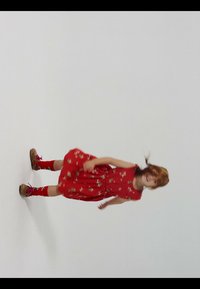 Ung pige med rødt hår iført en rød blomstret kjole, røde sokker og brune sko, der står mod en ensfarvet hvid baggrund og smiler.