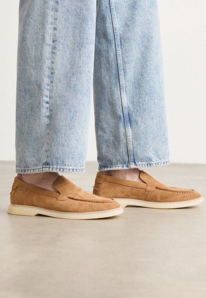 Des chaussures en suède brun à enfiler, à profil bas, avec une semelle extérieure crème et des détails de couture, assorties à un pantalon en denim bleu clair.