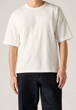 Mand iført almindelig hvid kortærmet t-shirt og mørkeblå jeans, stående med armene afslappet langs siden mod en neutral baggrund.