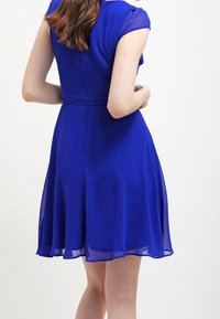 En kortärmad klänning i knälängd i livlig blå färg, med en figursydd kropp och en utsvängd kjol i ett svävande, lätt tyg.