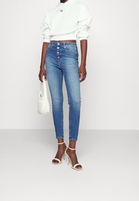 Smal blå denimjeans med hög midja och knappstängning fram, som matchas med en vit kort hoodie och beige sandaler med remmar.