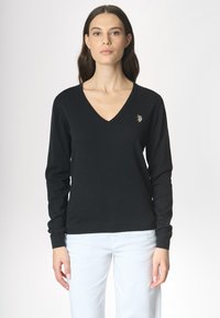 Una donna con capelli lunghi indossa un maglione nero a V, con un piccolo emblema, abbinato a pantaloni bianchi su uno sfondo chiaro.