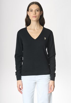 Una donna con capelli lunghi indossa un maglione nero a V, con un piccolo emblema, abbinato a pantaloni bianchi su uno sfondo chiaro.