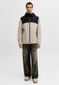 Veste bicolore beige et noire, avec fermeture éclair à l'avant et capuche. Caractérisée par un tissu léger et lisse et une coupe décontractée. Portée avec un jean large foncé.