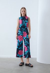 Črna brezrokavnik jumpsuit z velikim rožnatim in modrim cvetličnim vzorcem. Elastičen pas, široke hlače, ki se dopolnjujejo z lesenimi platformami.
