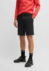 Pantaloni corti neri con una vestibilità standard, caratterizzati da una texture liscia e cinque tasche, abbinati a una felpa rossa brillante e scarpe da ginnastica nere.