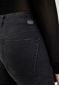 Jeans de mezclilla negra de corte ajustado. Las características incluyen un bolsillo trasero visible, costuras en contraste y una etiqueta de logo metálica.
