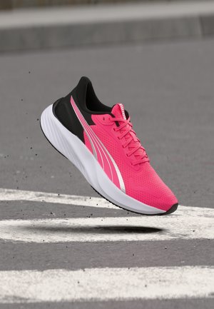 Élénk rózsaszín futócipő fekete sarokkal, fehér talppal és ezüst csíkkal, levegőben, egy szürke úton fehér jelzésekkel.