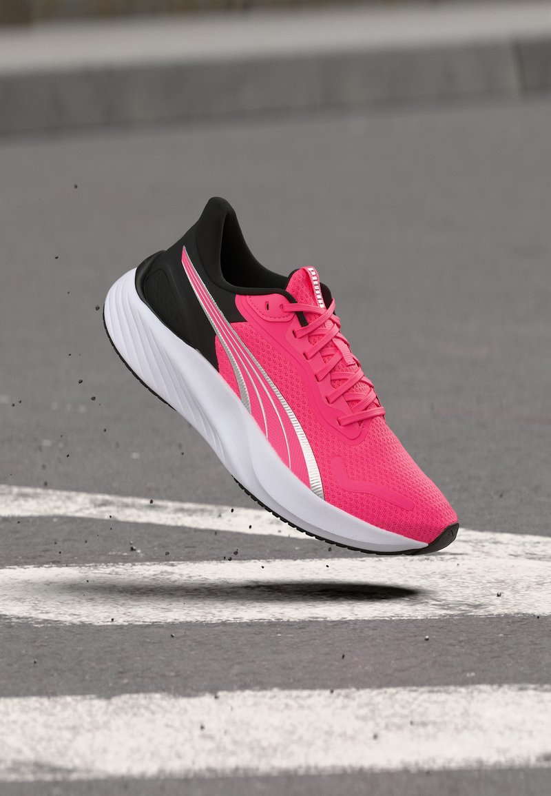 Φωτεινό ροζ παπούτσι τρεξίματος με μαύρη φτέρνα, λευκή σόλα και ασημένια λωρίδα, αιωρούμενο πάνω από έναν γκρι δρόμο με λευκές γραμμές.