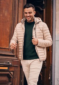 Beige pufferjack met een capuchon, ritssluiting aan de voorkant en horizontale stiksels, gedragen over een donkergroen ribbeltrui en lichte broek.