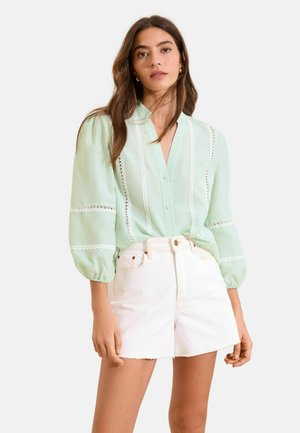 Frau trägt hellgrüne, gestreifte Bluse mit Spitzen-Details und weiße Denim-Shorts mit hoher Taille, steht mit entspannt an den Seiten hängenden Armen.