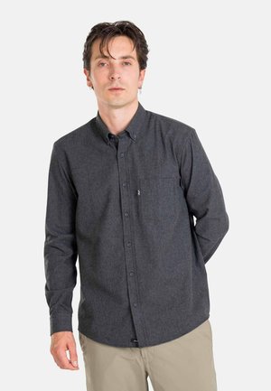 Man with dark hair wearing a dark gray button-up shirt and light beige pants standing against a white background.