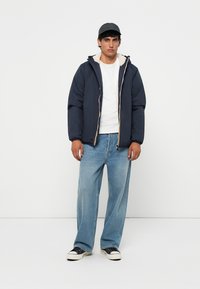 Padded navy jacket with a hood and white shearling lining, paired with light blue jeans and black sneakers. Zipper front and elastic cuffs.