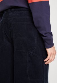 Pantalones de pana azul marino con textura acanalada, que cuentan con dos bolsillos traseros y un bolsillo lateral; combinados con una camisa de manga larga morada.