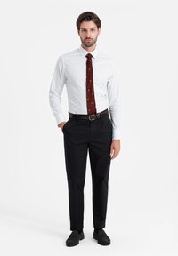Camisa blanca de manga larga con pequeños lunares azules marinos, combinada con una corbata burdeos con patrones claros y pantalones negros. Zapatos negros completan el look.