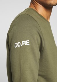 Olivgrön långärmad skjorta med ribbad textur och rundad halsringning. Har vit "CO.RE"-text på vänster ärm.