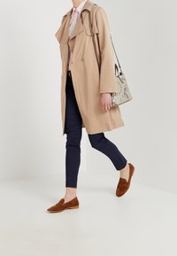 Beige trenchcoat med stora slag, ljusrosa skjorta, marinblå slim-fit byxor och bruna mockaloafers, i kombination med en mönstrad axelväska.