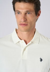 Polo shirt beige chiaro realizzato in cotone, con colletto classico, tre bottoni e un piccolo logo navy ricamato sul lato sinistro del petto.
