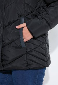 Chaqueta de plumas negra con un patrón acolchado, que presenta un bolsillo lateral resaltado por un acento gris. La tela parece suave y brillante.