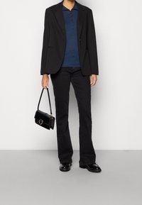 Svart blazer över en marinblå polo, matchad med svarta utsvängda jeans. Tillbehören inkluderar en liten svart handväska med gulddetaljer och skinande svarta skor.