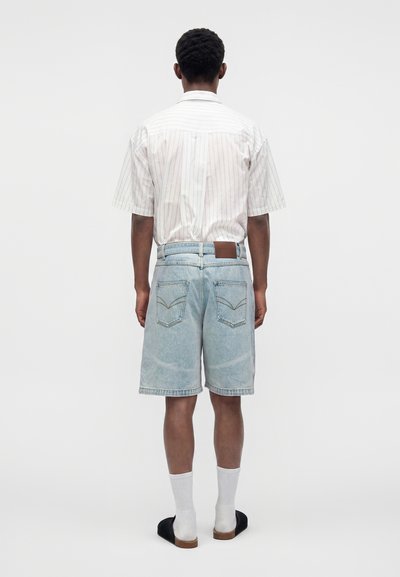 Homme de dos portant une chemise blanche rayée à manches courtes, un short en jean bleu clair, des chaussettes blanches et des chaussures noires à talons ouverts.