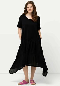 Schwarzes, gestuftes Kleid mit kurzen Ärmeln und V-Ausschnitt. Verfügt über Taschen und einen längeren Saum hinten. Getragen mit pinken Sandalen.