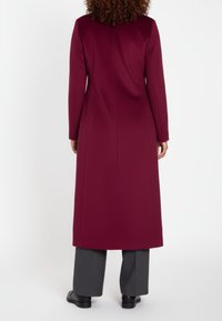 Bordeauxroter langer Mantel mit geradem Schnitt, taillierten Nähten und langen Ärmeln, kombiniert mit grauen Hosen und schwarzen Schuhen.