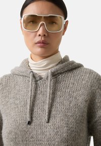Close-up van een persoon die een oversized witte bril met getinte glazen draagt en een grijze gebreide hoodie over een witte turtleneck heeft, tegen een effen achtergrond.