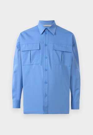 Camisa de manga longa azul clara com gola pontiaguda, frente com botões e dois grandes bolsos de aba no peito, exibida sobre um fundo branco.