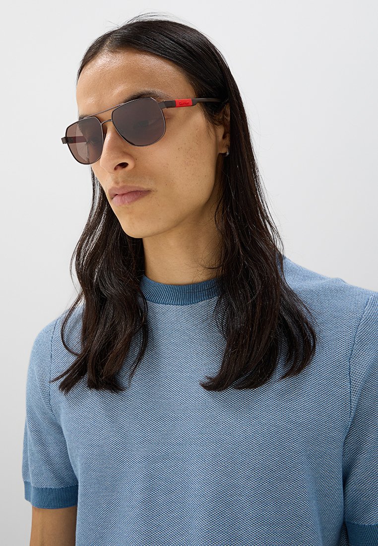 Gafas de sol con lentes oscuros y un delgado marco metálico negro. Acentos rojos en las patillas. Llevadas con una camiseta de punto texturizado azul.