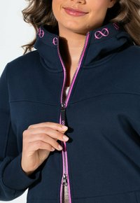 Donna che indossa una felpa con zip blu navy con rifiniture rosa e simboli dell'infinito rosa sul tirazip e sul colletto, tenendo la zip a metà.