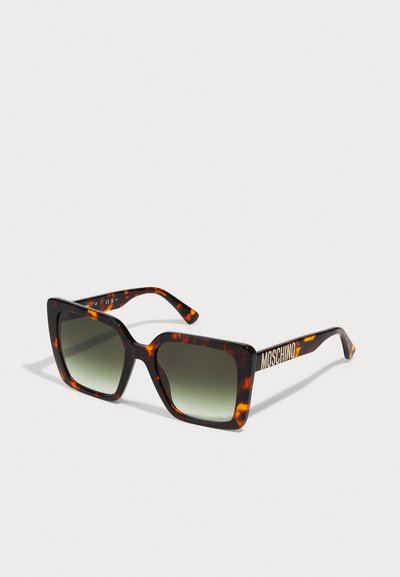 Ochelari de soare tip țestoasă cu un cadru pătrat larg, lentile degradé gri-verde și logo-ul auriu "Moschino" pe braț.