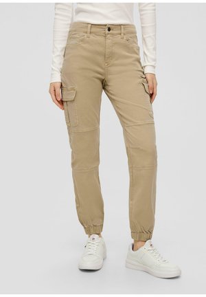 Beige cargobroek met elastische boorden, meerdere zakken en een gladde textuur. Gecombineerd met witte sneakers voor contrast.
