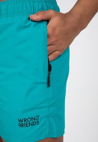 Pantaloni da bagno turchesi con tasca con zip, dotati di vita elasticizzata e logo stampato "WRONG FRIENDS" sulla gamba sinistra.