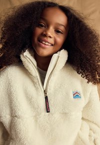 Pull en polaire blanche avec un col zippé, arborant un écusson bleu et orange sur la poitrine. Texture tactile et douce avec un design confortable.