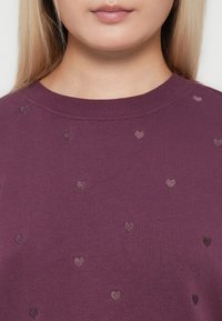 Närbild av en person som bär en lila tröja med små broderade hjärtmönster nära halsringningen.