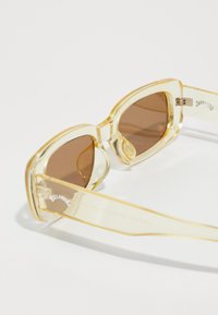 Gafas de sol translúcidas amarillas con lentes marrón ahumado, que presentan un diseño distintivo y un sutil detalle de logotipo en la patilla.