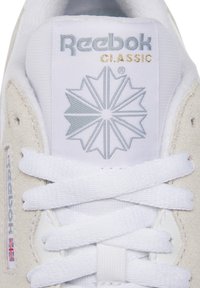 Close-up valge Reebok Classic kinga keel hõbedase logoga, kuldse "CLASSIC" tekstiga, valgete paeltega ja sametise külgpaneeliga, millel on Briti lipu silt.