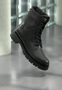 Μαύρο δερμάτινο μποτάκι μάχης με κορδόνια και παχιά ραφτή σόλα που αιωρείται πάνω σε μια λεία γκρι επιφάνεια σε έναν σύγχρονο εσωτερικό χώρο.