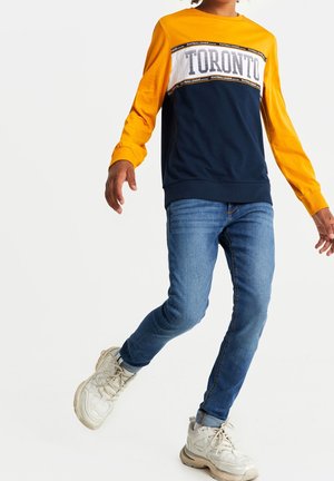 Sudadera de manga larga con mangas amarillas y cuerpo azul marino, con un panel blanco en el pecho con el texto "Toronto"; combinado con jeans azules y zapatillas.