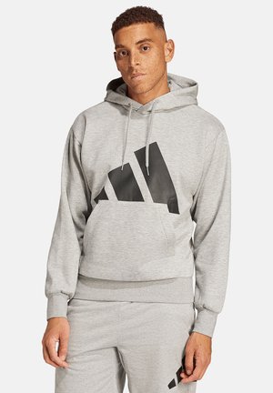 Man with short curly hair wearing light gray hoodie and matching sweatpants with large black Adidas logo on hoodie front.