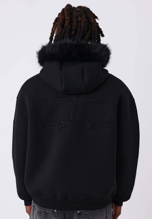 Personne aux cheveux bruns torsadés porte un sweat à capuche noir avec une bordure en fourrure sur la capuche et un logo embossé "PXFP" dans le dos, dos à la caméra.