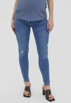 Persoon in blauwe skinny jeans met lichte beschadigingen en donkere open teen hakken, staand tegen een effen achtergrond.
