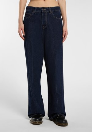 Personne portant un jean bleu foncé à jambes larges et des chaussures noires, debout devant un fond clair uni.
