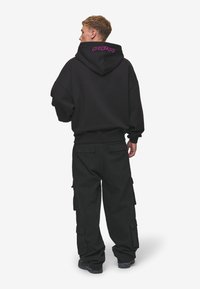 Čierna mikina s kapucňou a ružovou vyšitou nášivkou na chrbte, kombinovaná s voľnými čiernymi cargo nohavicami so bočnými vreckami.