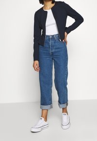 Marineblauwe cropped jack, witte getailleerde t-shirt, lichtblauwe high-waisted jeans, opgerolde mouwen, en witte sneakers met zwarte accenten.
