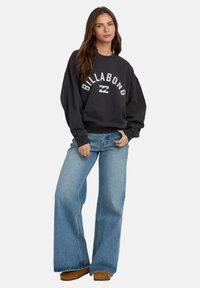Czarny sweter z logo "BILLABONG", szerokimi rękawami i ściągaczami, zestawiony z jasnoniebieskimi dżinsami w kształcie dzwonów oraz brązowymi butami wsuwanymi.
