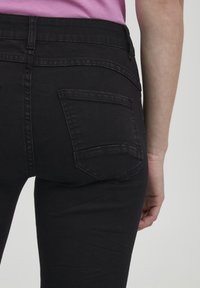 Jeans de mezclilla negra con un corte ajustado, que cuentan con bolsillos traseros y una textura suave. La cintura es visible con un ligero desgaste.
