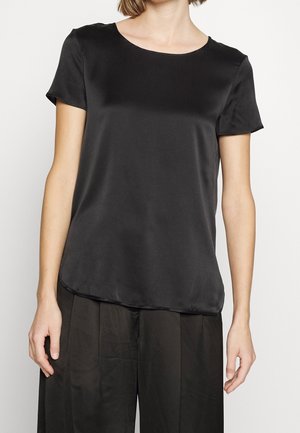 Kobieta ubrana w prostą czarną satynową bluzkę z krótkim rękawem i luźne czarne spodnie, stojąca na jasnym tle.